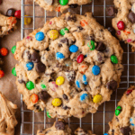 A large, chunky cookie filled with colorful candy-coated chocolates and chocolate chips sits on a cooling rack, surrounded by other similar cookies.
