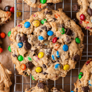 A large, chunky cookie filled with colorful candy-coated chocolates and chocolate chips sits on a cooling rack, surrounded by other similar cookies.
