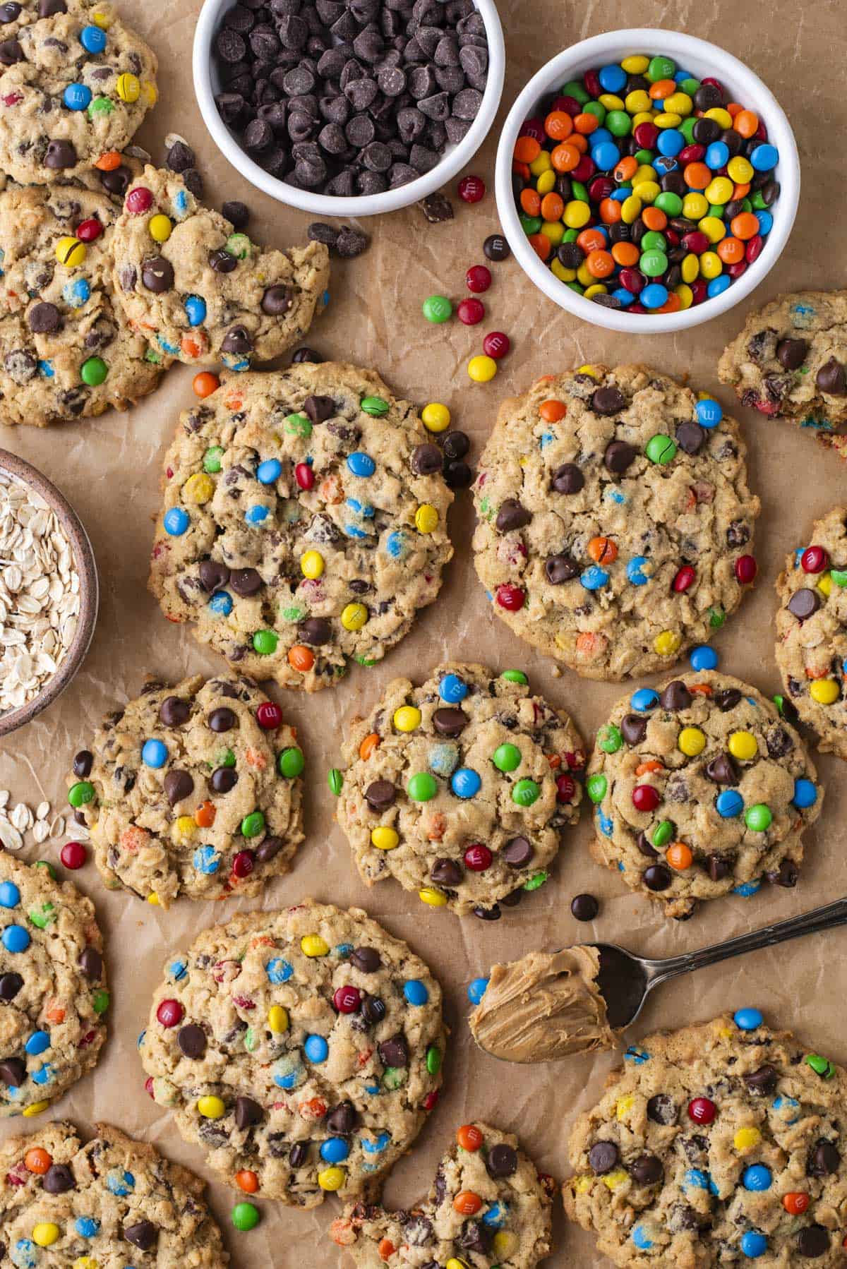 A batch of large cookies with colorful chocolate candies and chocolate chips sits on parchment paper. Nearby are bowls of chocolate chips, colorful candies, oats, and a spoonful of peanut butter.