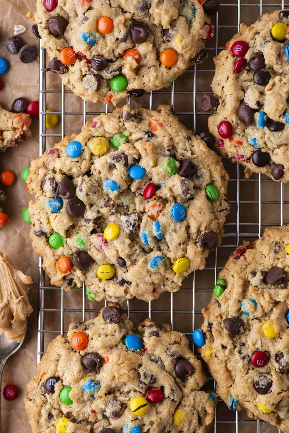 Large cookies filled with colorful candy-coated chocolates and chocolate chips rest on a cooling rack. A spoonful of peanut butter and cookie crumbs are visible on brown parchment paper nearby.