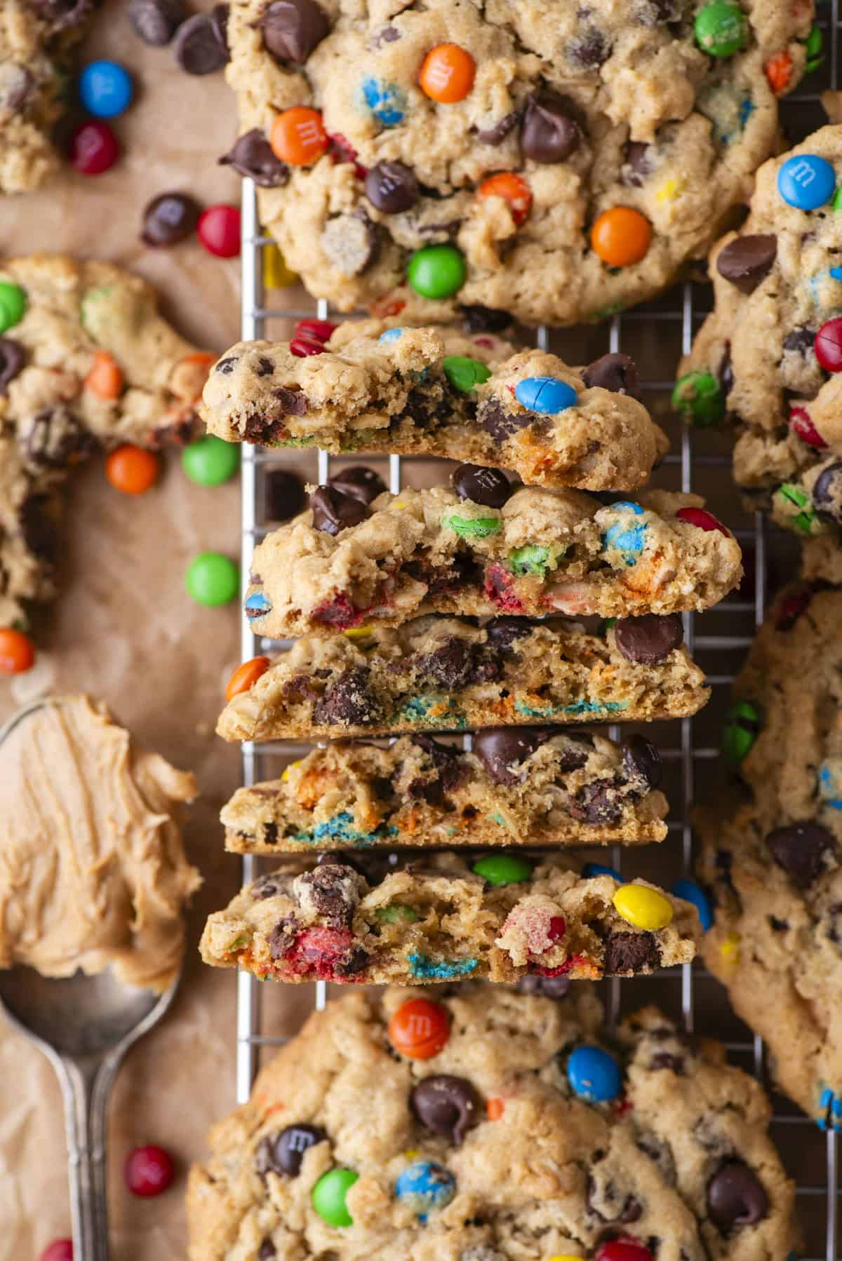 A stack of thick, chewy cookies filled with colorful chocolate candies and chocolate chips, broken in half to show their soft texture, sits on a cooling rack with a spoonful of peanut butter nearby.