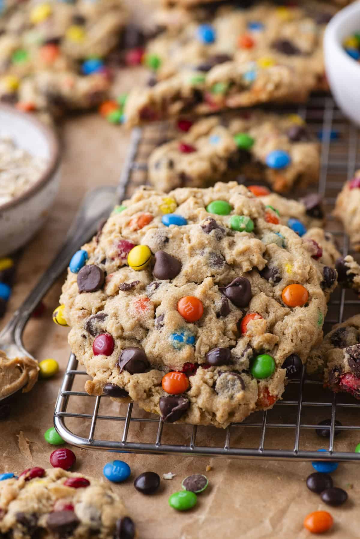 A close-up of colorful cookies with M&M candies and chocolate chips cooling on a wire rack, with more cookies and scattered candies in the background.