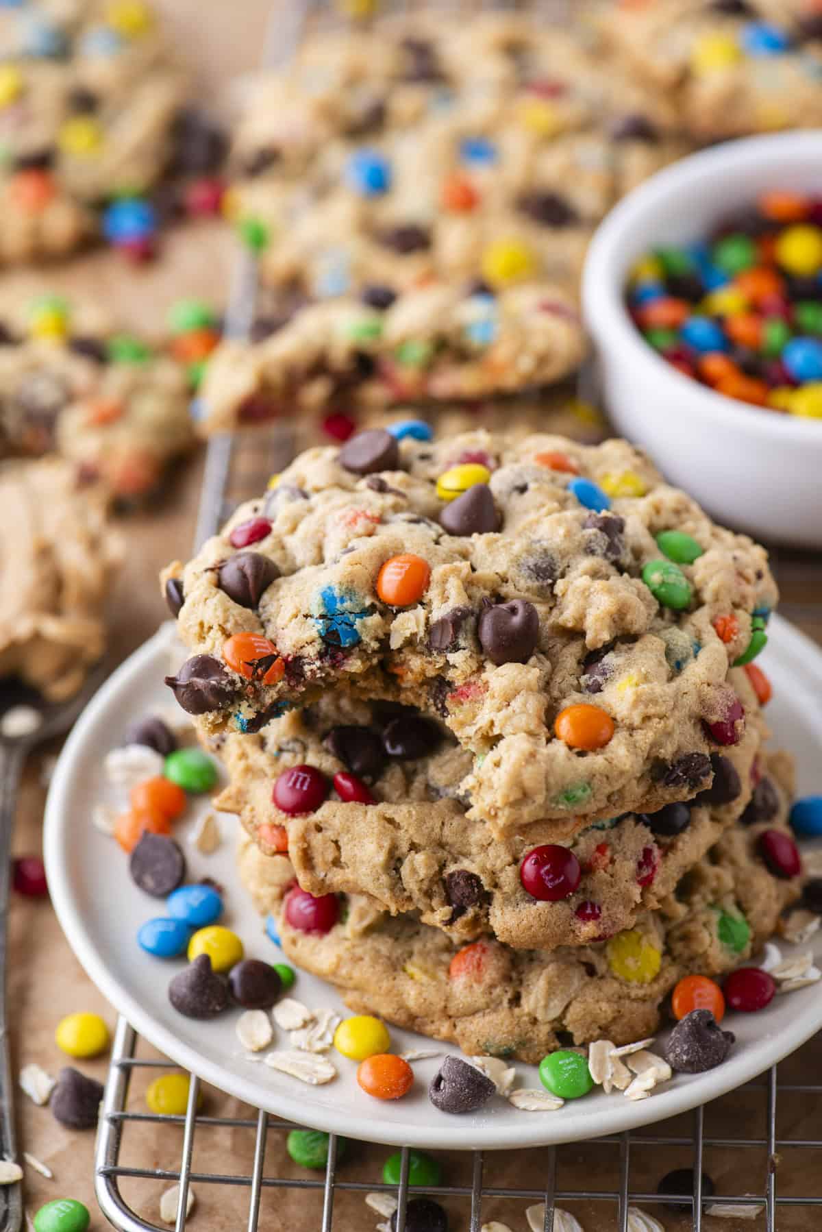 A stack of colorful cookies with chocolate chips and candy-coated chocolates sits on a white plate, with one cookie on top showing a bite taken out. More cookies and a bowl of candies are in the background.