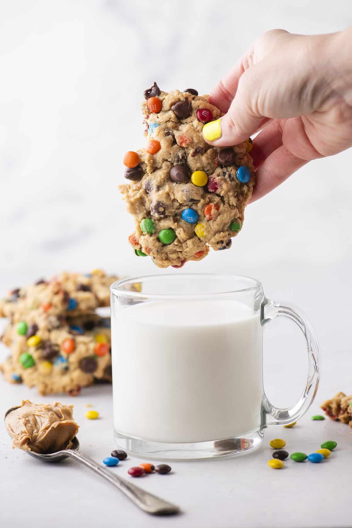 A hand dips a colorful candy-studded cookie into a clear glass mug of milk. More cookies, a spoon with peanut butter, and scattered candies are on the white surface nearby.