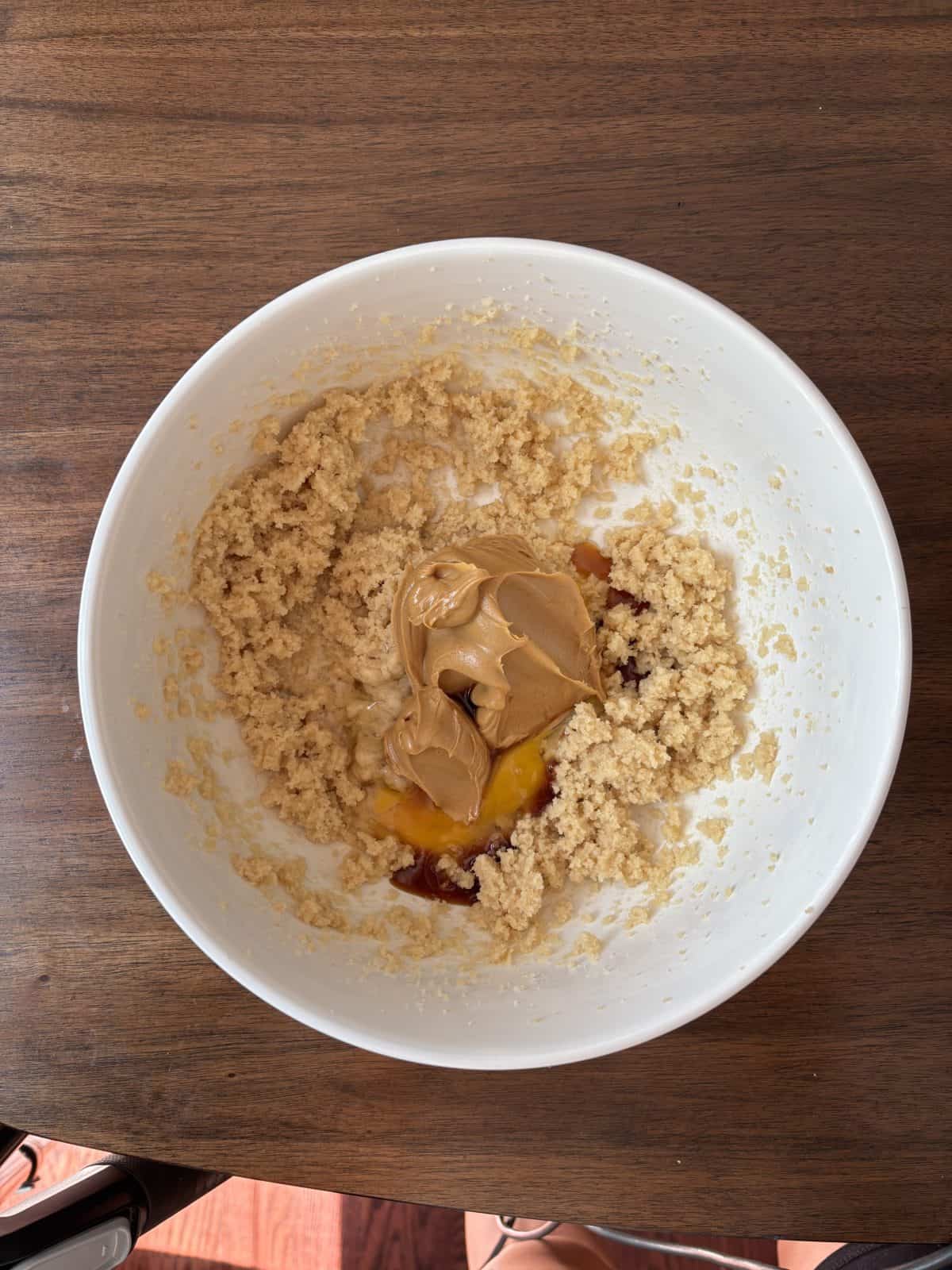 A white bowl on a wooden table contains a mixture of creamed sugar and butter, with peanut butter, egg, and vanilla extract added but not yet mixed in.