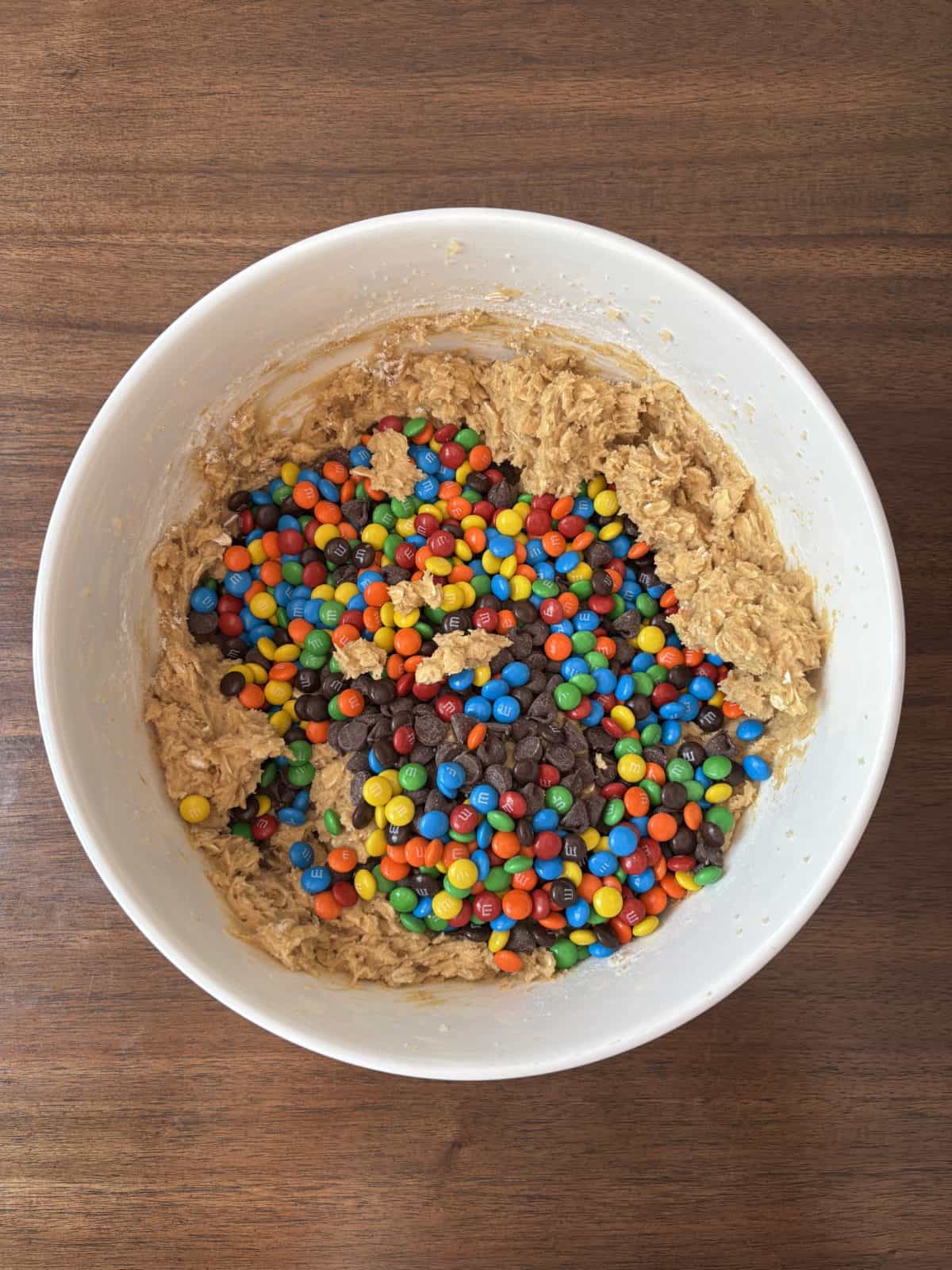 A white mixing bowl on a wooden surface containing cookie dough mixed with colorful mini chocolate candies and chocolate chips.