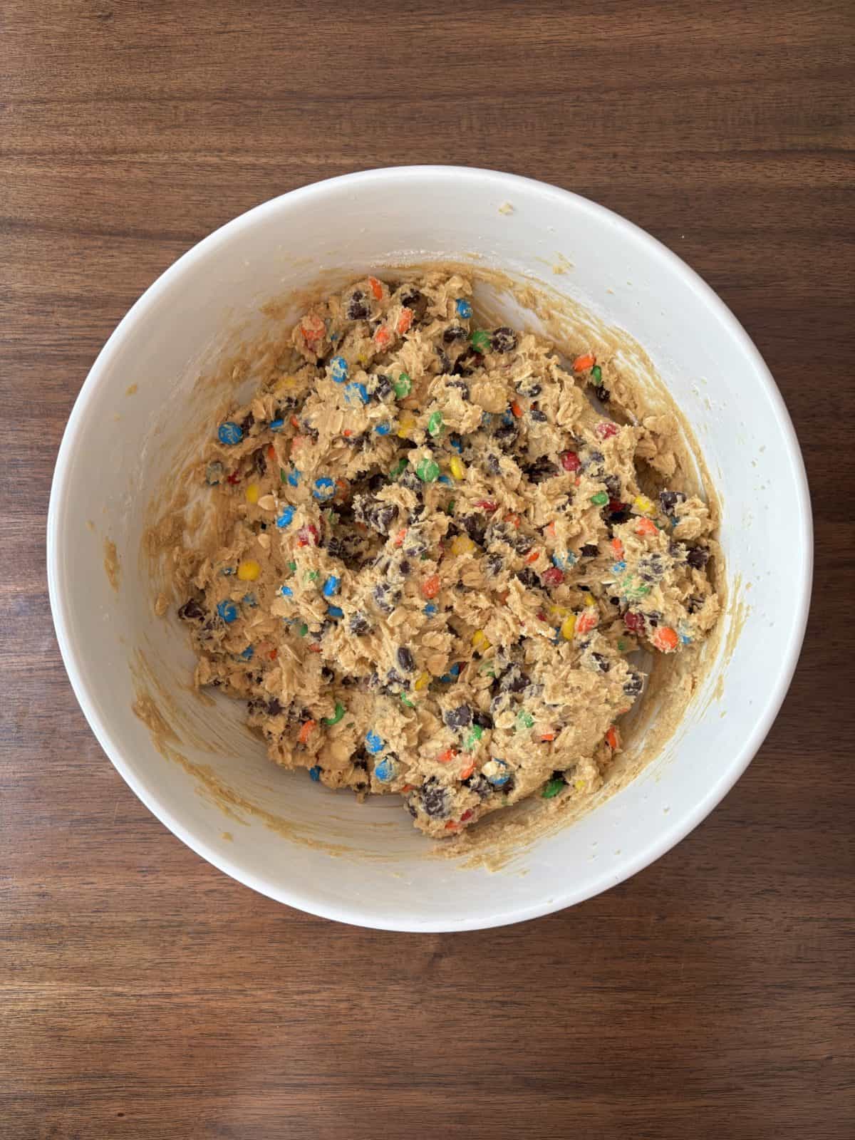 A white bowl filled with cookie dough mixed with chocolate chips and colorful candy pieces sits on a wooden surface.
