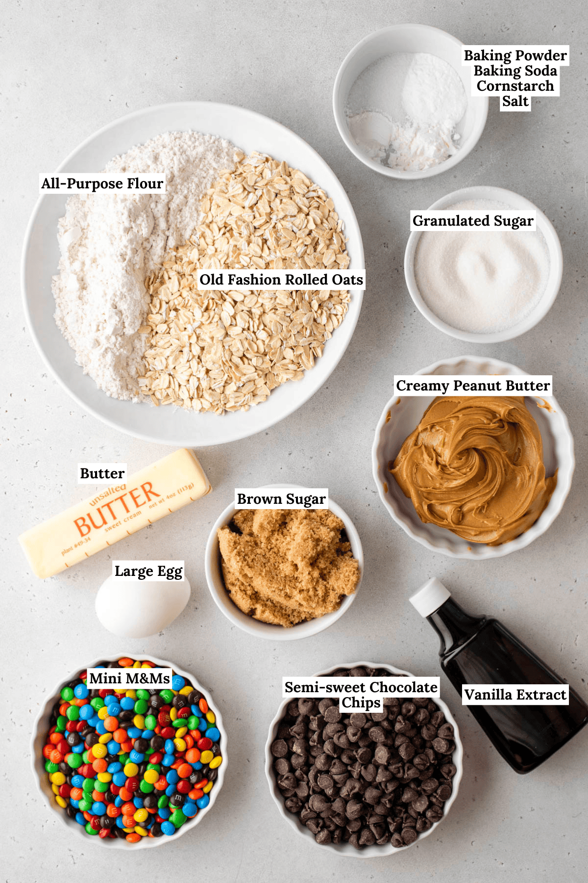A top-down view of labeled ingredients for cookies: flour, rolled oats, granulated sugar, brown sugar, butter, egg, creamy peanut butter, baking powder, baking soda, cornstarch, salt, vanilla, chocolate chips, and mini M&Ms.