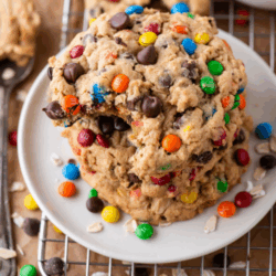 A stack of chunky monster cookies with chocolate chips and colorful candy-coated chocolates sits on a white plate, with a bite taken from the top cookie. Scattered candies and oats are visible around the plate.