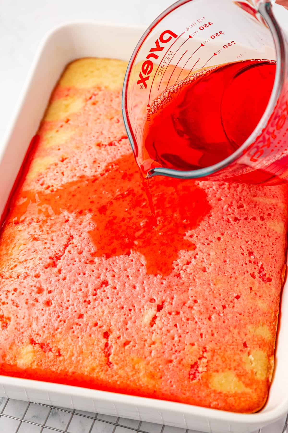 A glass measuring cup pours red liquid over a rectangular yellow cake in a white baking dish, with the cake showing small holes where the liquid is being absorbed.