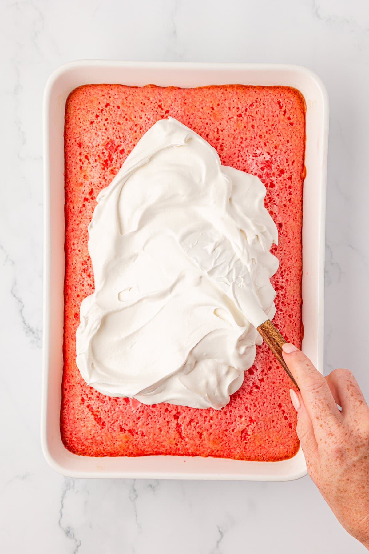 A hand spreads a thick layer of white whipped topping over a rectangular pink cake in a white baking dish on a marble surface.