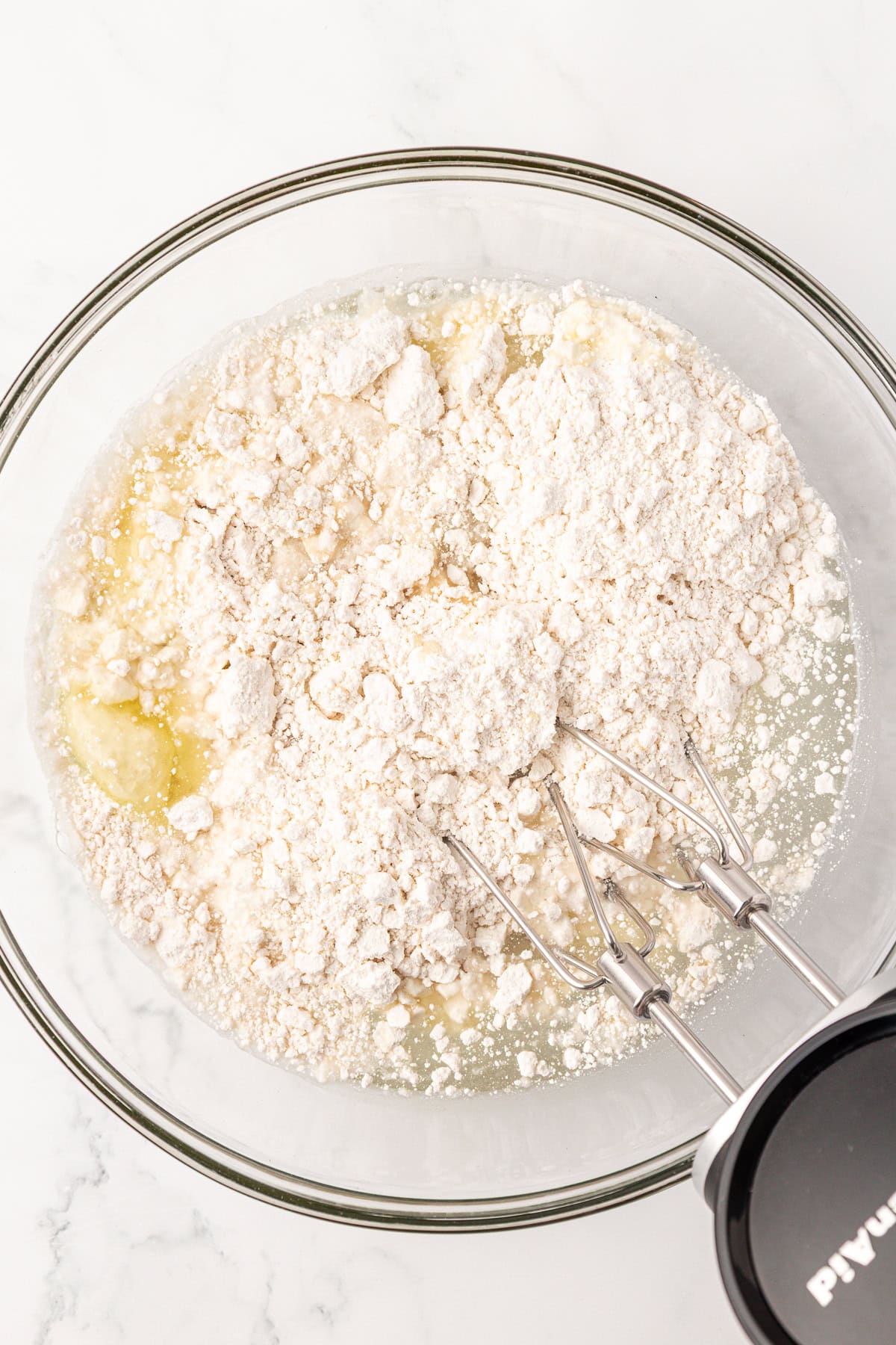 A glass bowl containing flour and wet ingredients is being mixed with an electric hand mixer on a white surface.