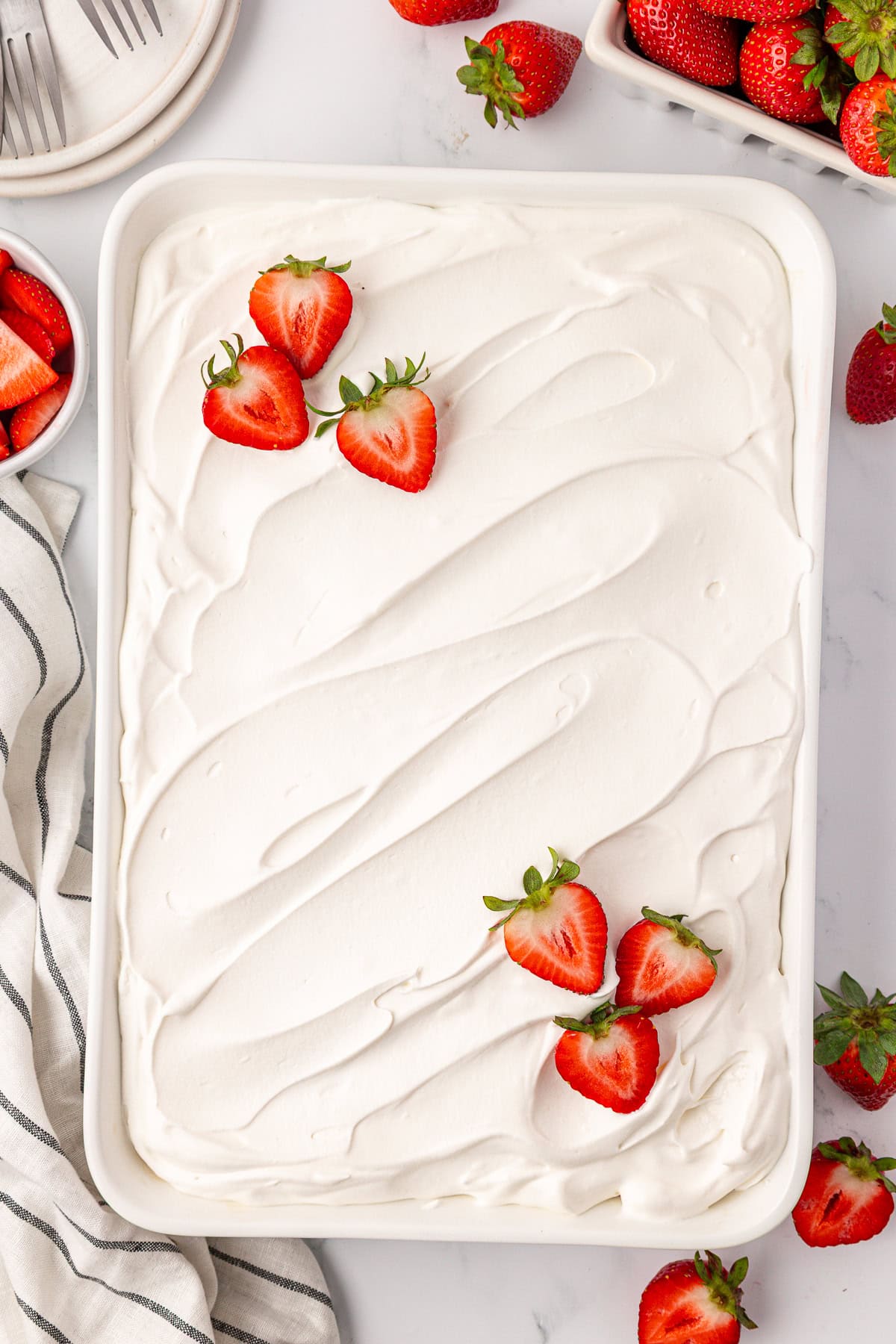 A rectangular dish filled with a dessert topped with smooth, white whipped cream, garnished with fresh, halved strawberries. Whole and sliced strawberries are scattered nearby on a white surface.