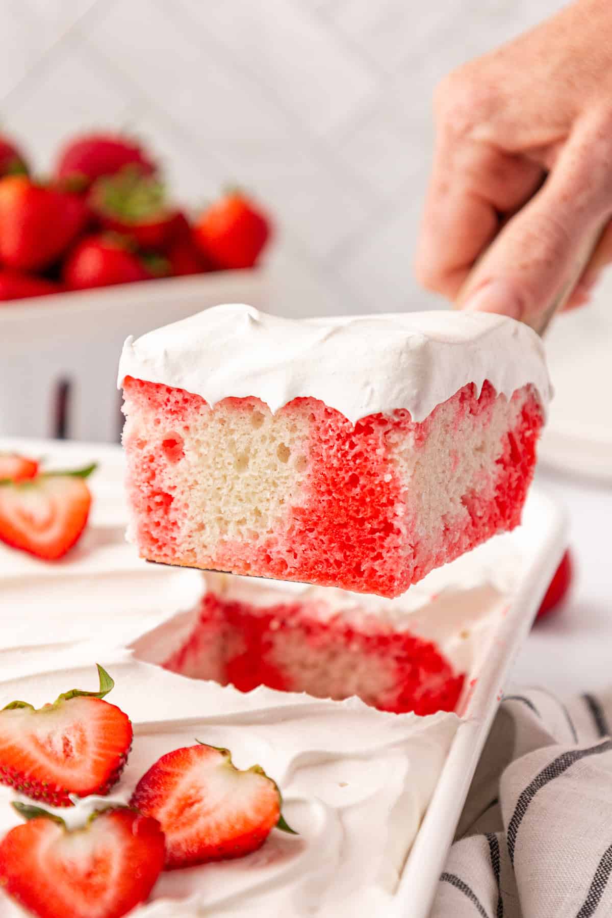 A hand lifts a slice of white and red poke cake topped with whipped cream from a tray. Fresh strawberries are on the tray and in a bowl in the background.
