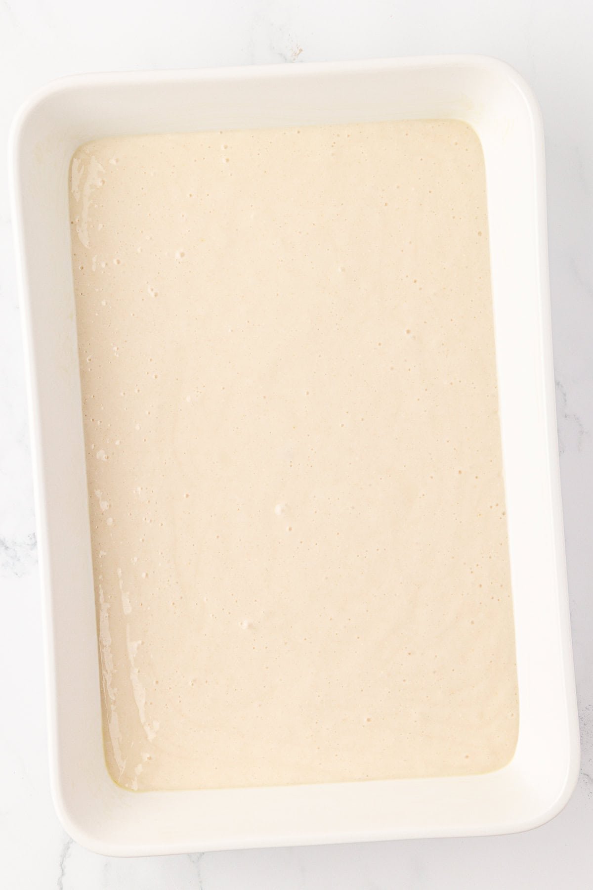 A white rectangular baking dish filled with an even layer of light beige cake batter, set on a white marble surface.