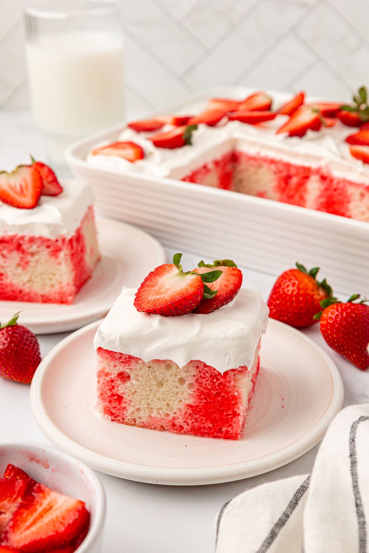 A slice of strawberry poke cake topped with whipped cream and fresh strawberry slices on a plate, with more cake and strawberries in the background.