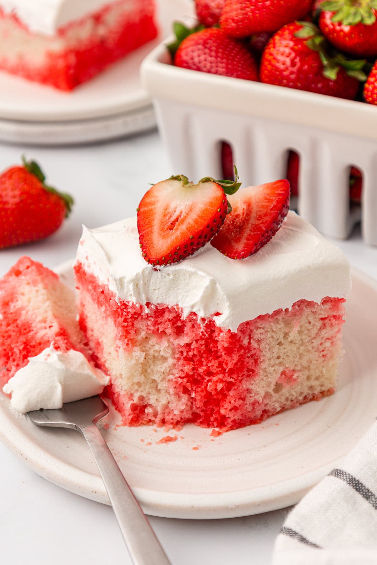 A slice of strawberry poke cake with white frosting and two halved strawberries on top sits on a plate with a fork. In the background, there is a container filled with fresh strawberries.