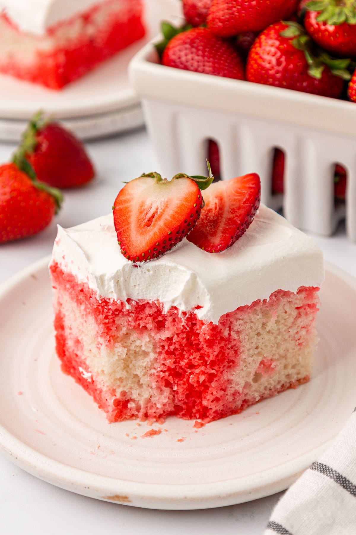 A slice of strawberry poke cake with white frosting and two strawberry halves on top, served on a plate with fresh strawberries and a container of strawberries in the background.