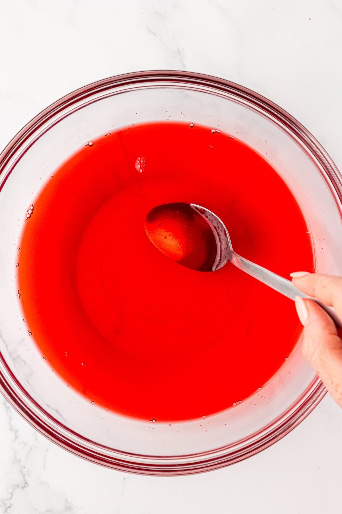 A hand holding a spoon stirs bright red liquid in a clear glass bowl on a white surface.