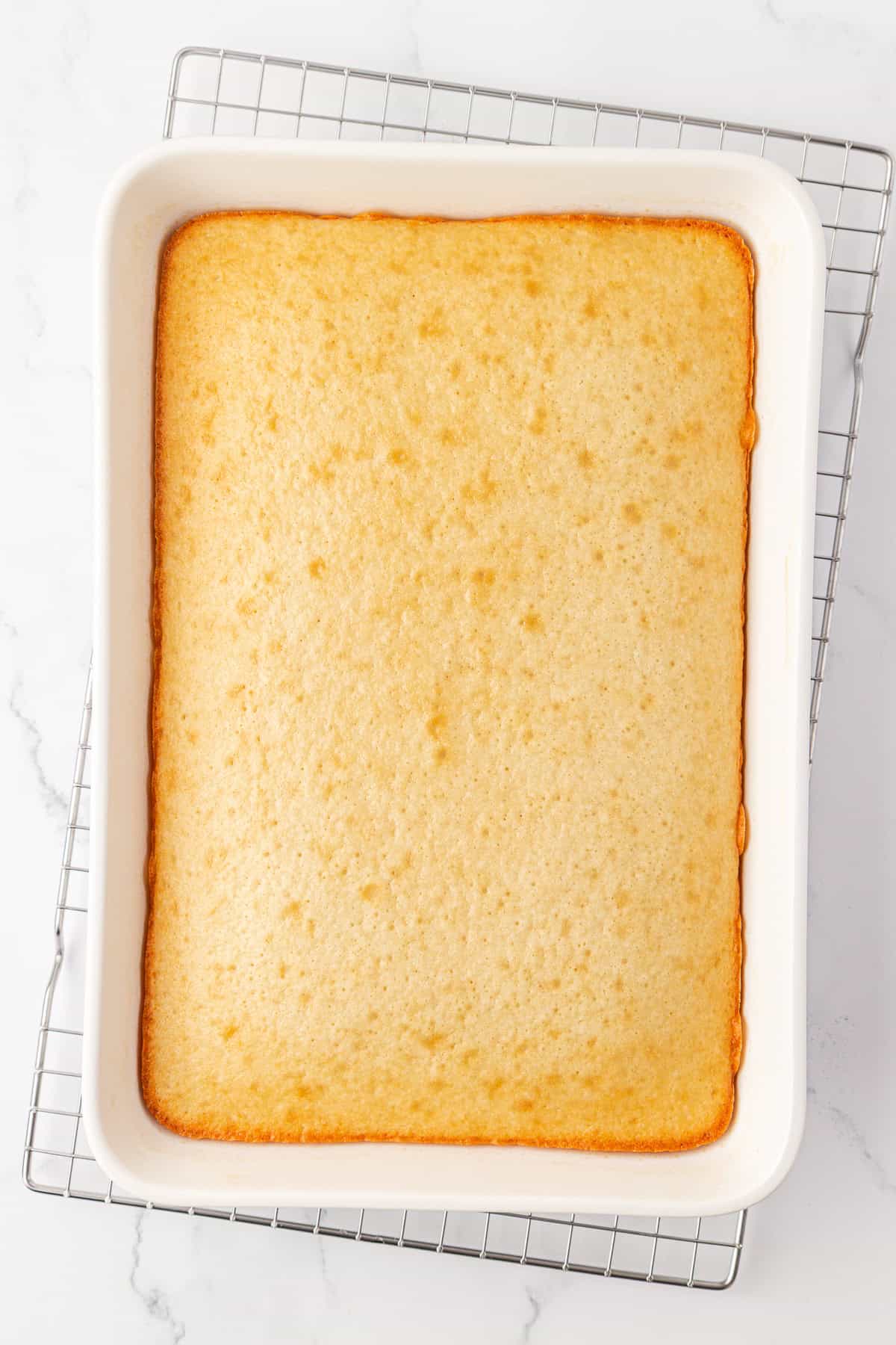 A rectangular vanilla sheet cake in a white baking dish, resting on a metal cooling rack atop a white marble surface. The cake has a golden-brown edge and a smooth, lightly textured top.
