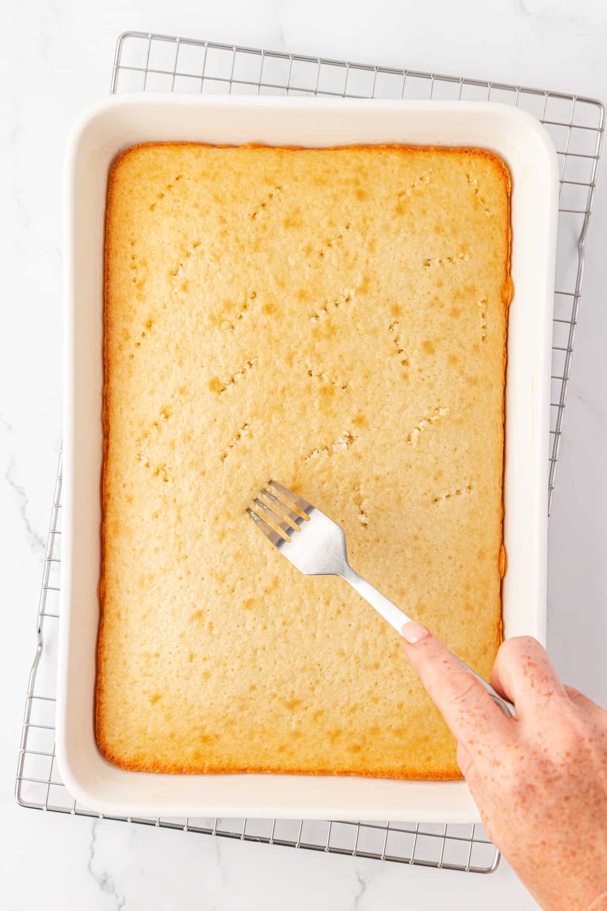 A hand uses a fork to poke holes in a rectangular, golden-brown cake still in a white baking dish, which rests on a metal cooling rack over a marble surface.
