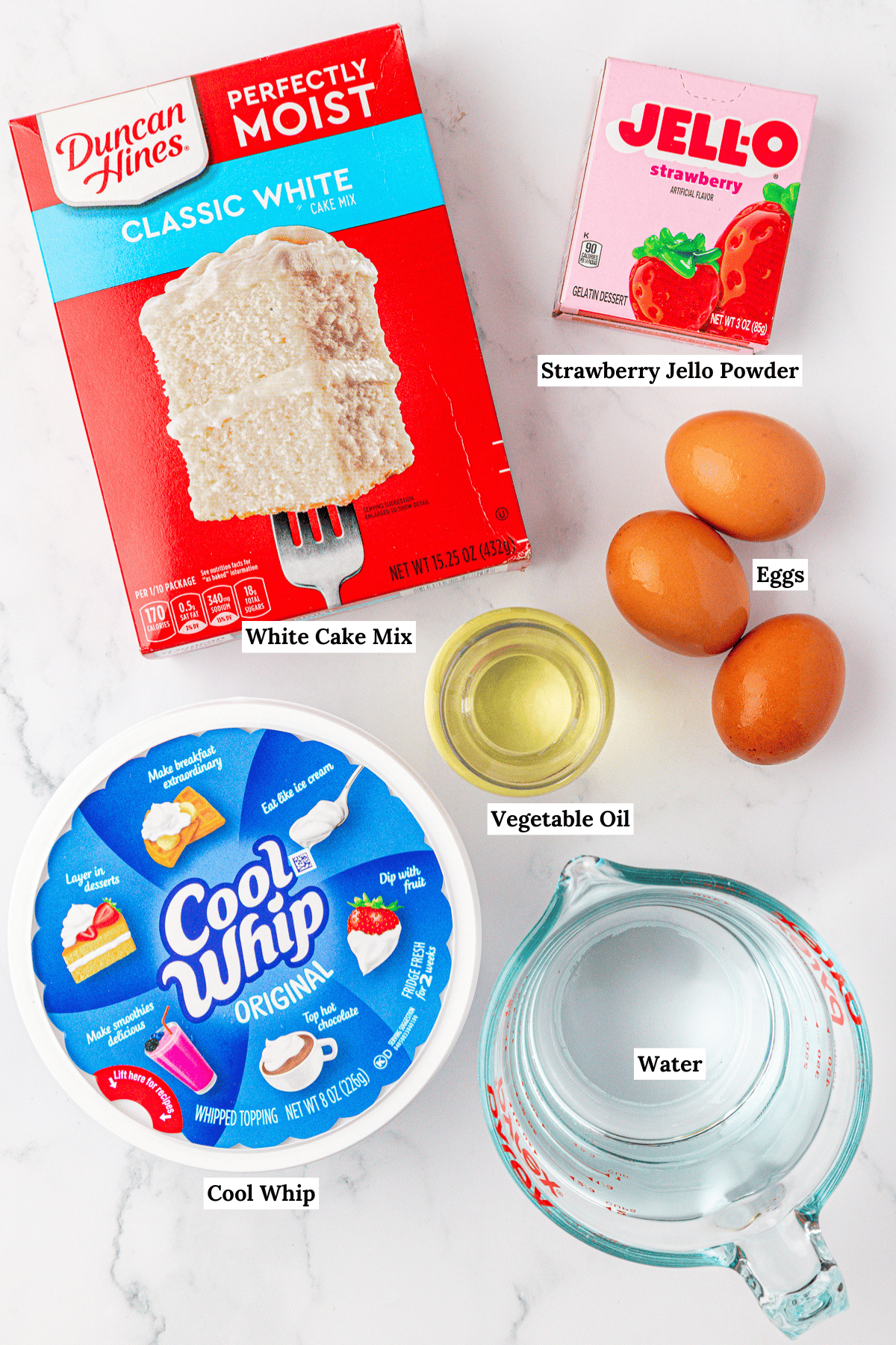 Top-down view of ingredients for a dessert: white cake mix, strawberry Jello powder, three eggs, vegetable oil in a small bowl, Cool Whip container, and a measuring cup of water on a white marble surface.