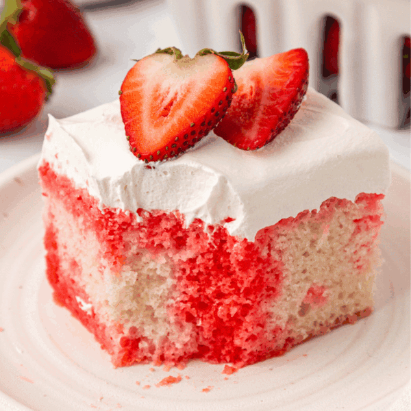 A slice of strawberry poke cake with white frosting, topped with two fresh strawberry halves, sits on a white plate. Fresh strawberries are visible in the background.