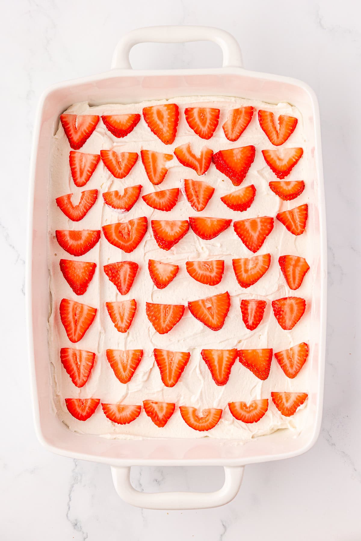 A white rectangular baking dish filled with a dessert topped with rows of halved fresh strawberries arranged neatly on a layer of white cream.