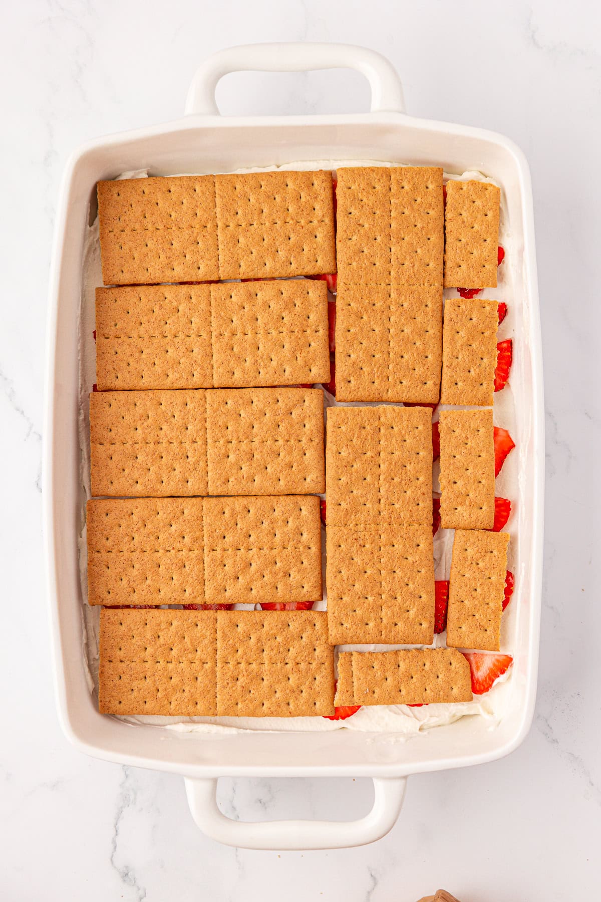 A white baking dish filled with a dessert layer of graham crackers on top of cream and sliced strawberries, sitting on a marble surface.