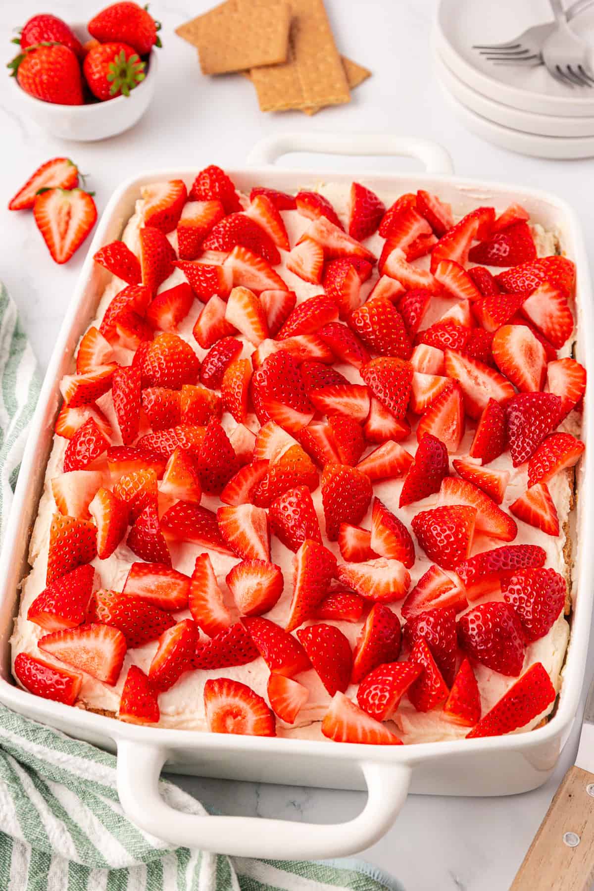 A white baking dish filled with a creamy dessert topped generously with sliced fresh strawberries. Nearby are plates with forks, whole strawberries, graham crackers, and a green-striped kitchen towel.
