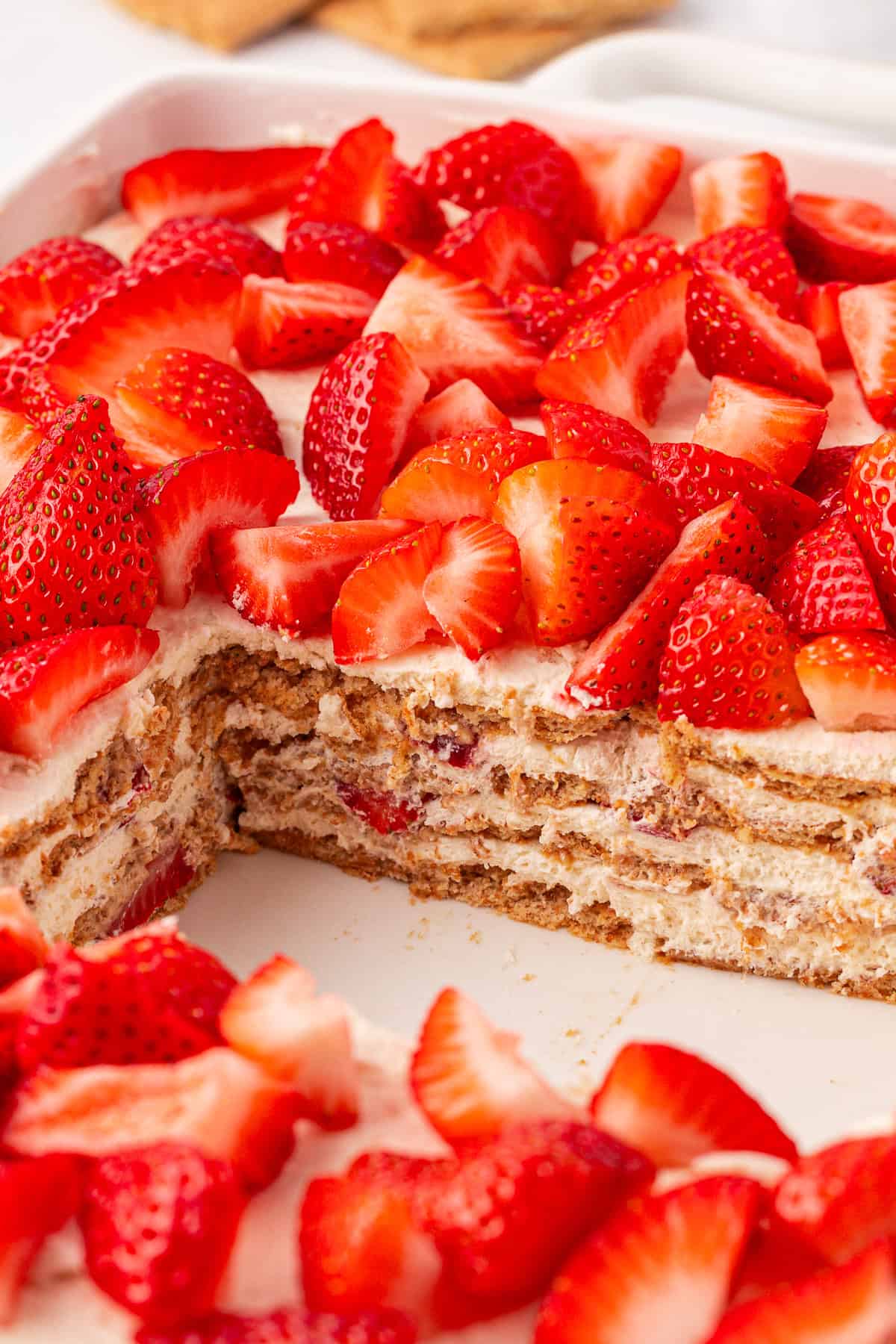 A layered dessert with cream, graham crackers, and fresh sliced strawberries, displayed in a dish with a large square piece removed to show the layers inside.