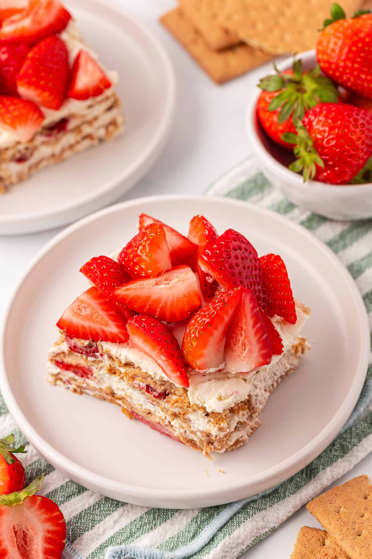 A slice of layered strawberry dessert with whipped cream and graham crackers on a white plate, topped with fresh strawberry pieces. Whole strawberries and graham crackers are in the background.