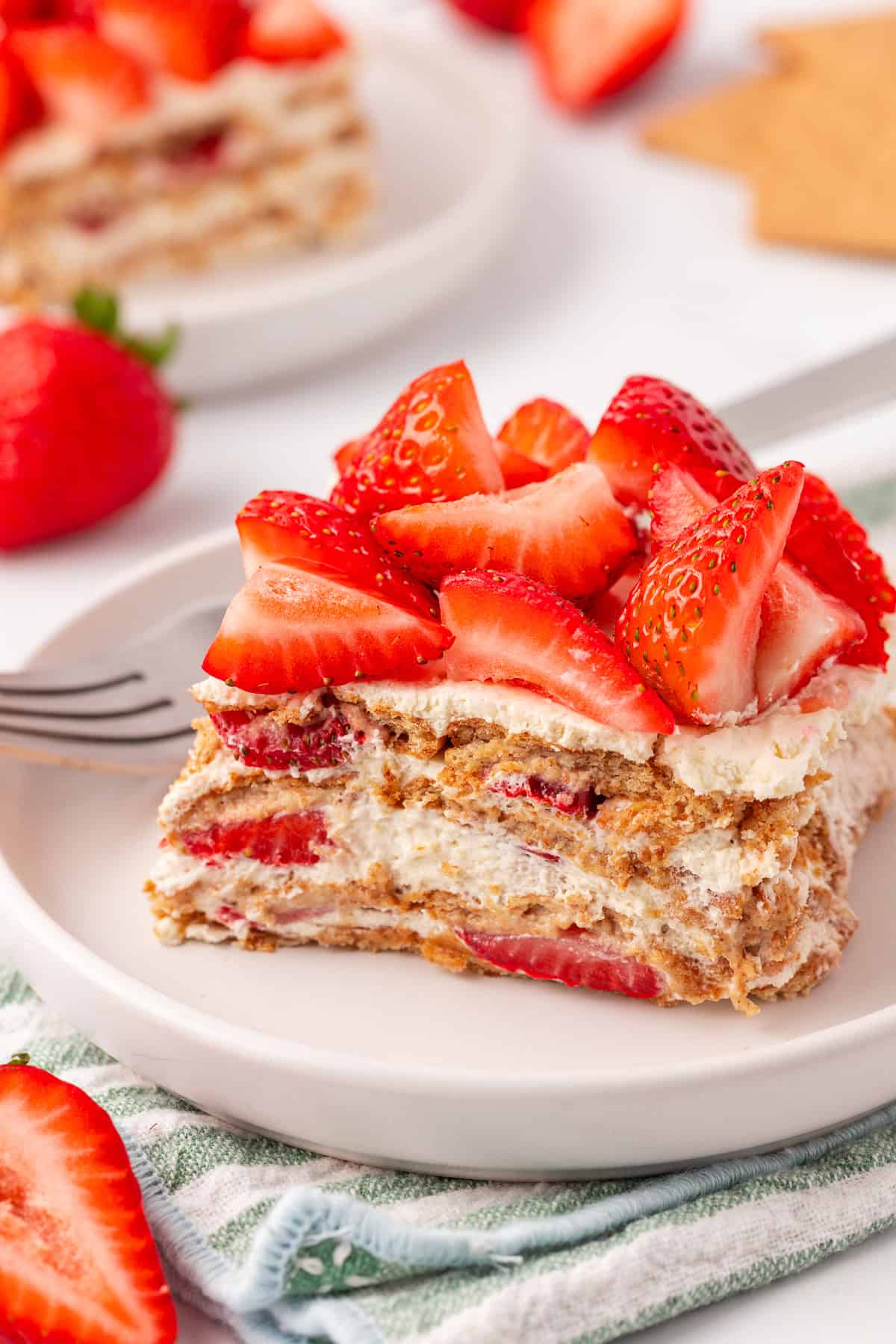 A slice of strawberry icebox cake with layers of whipped cream, graham crackers, and fresh strawberries on a white plate, garnished with strawberry pieces. A fork and additional strawberries are nearby.
