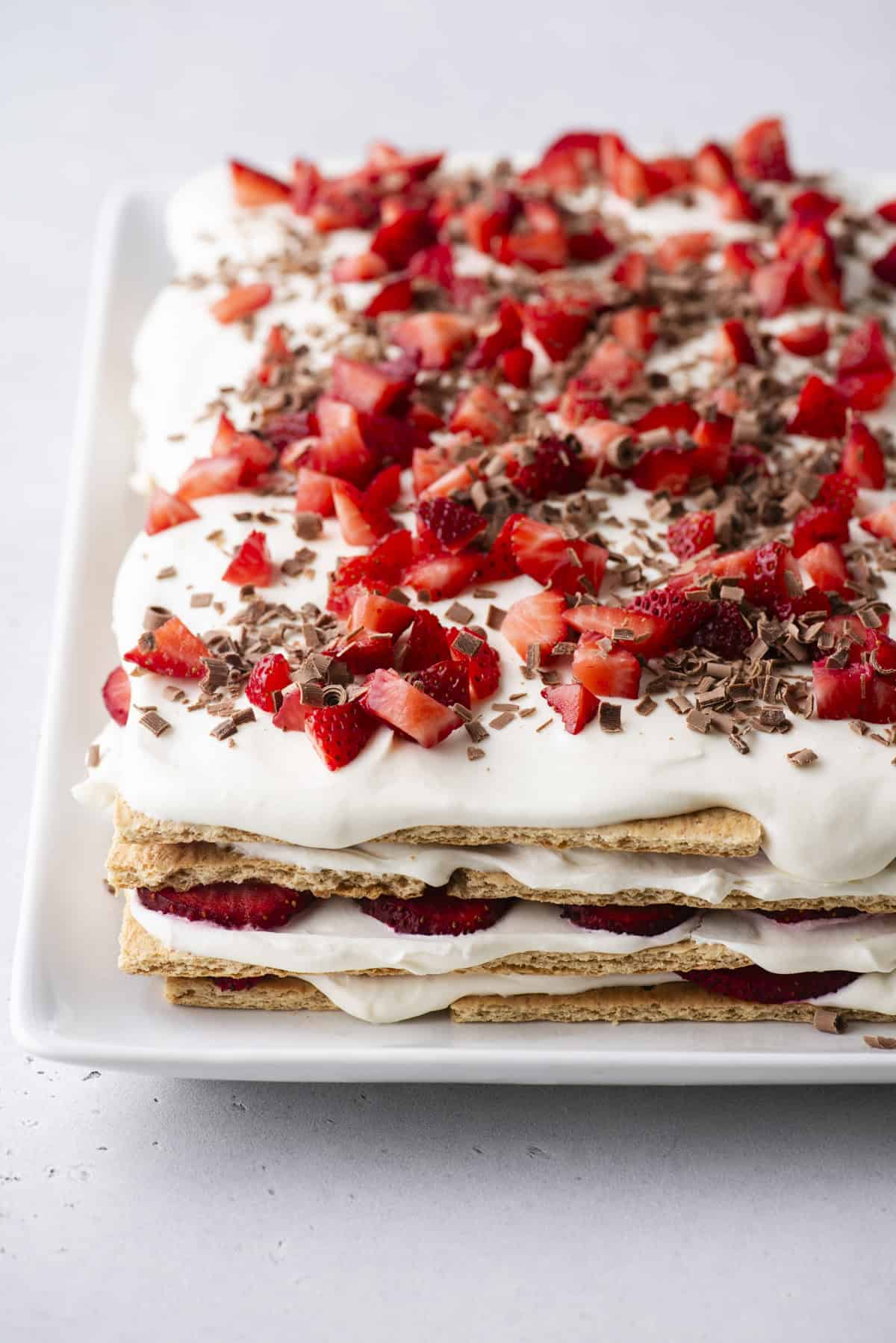 A layered dessert made with graham crackers, whipped cream, sliced strawberries, and chocolate shavings on a white rectangular plate.