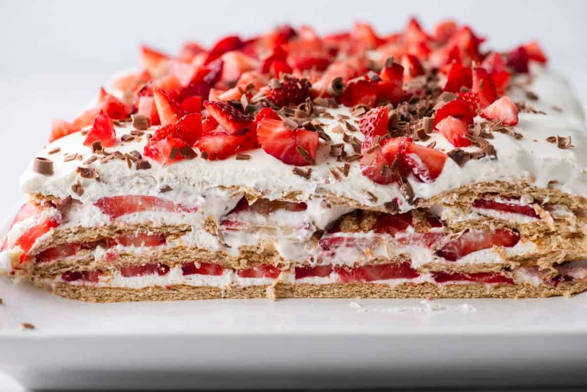 A rectangular layered dessert with whipped cream, sliced strawberries, graham crackers, and chocolate shavings, shown with a slice removed to reveal the layers inside.