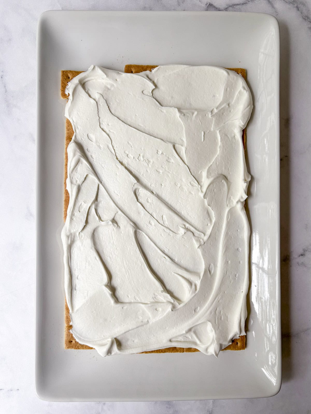 A rectangular graham cracker topped with a thick, even layer of white frosting sits on a white rectangular plate against a marble background.