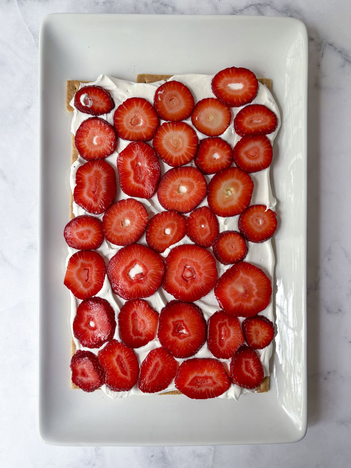 Rectangular dessert with a layer of whipped cream topped with neatly arranged fresh strawberry slices on a rectangular white plate, set on a light marble surface.