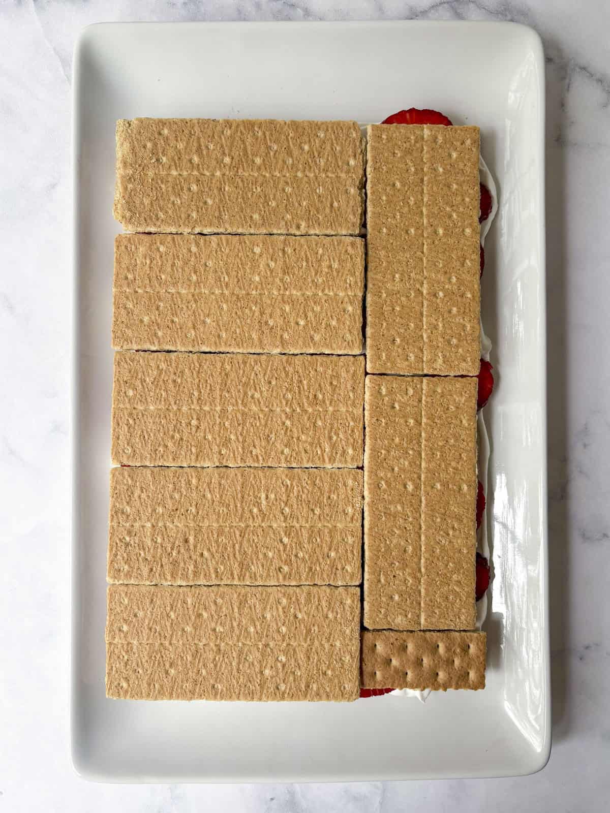 A rectangular white tray filled with neatly arranged graham cracker sheets covering a dessert with visible layers of strawberries and cream peeking out from the side.