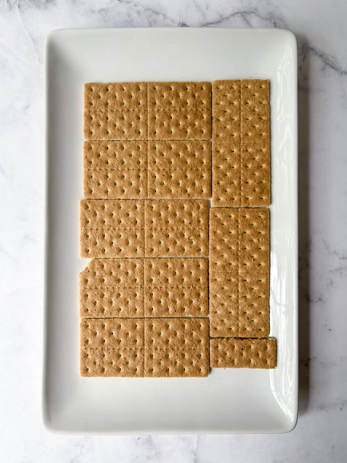 A white rectangular plate holds 14 rectangular graham crackers neatly arranged, with one cracker missing a corner and one small, separate square piece on the bottom right. The background is a light marble surface.