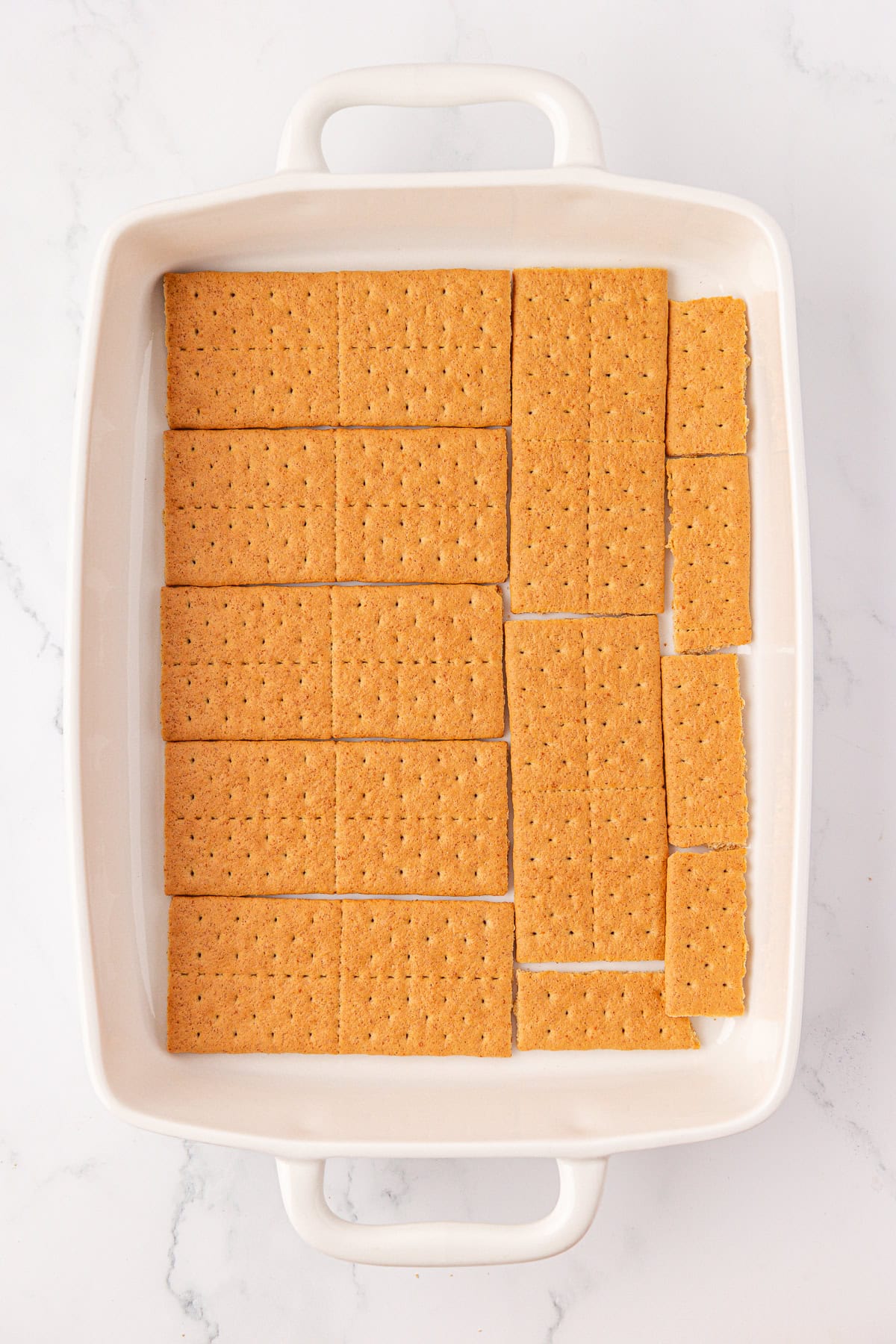 A white rectangular baking dish lined with pieces of graham crackers arranged to cover the bottom, set on a white marble surface.