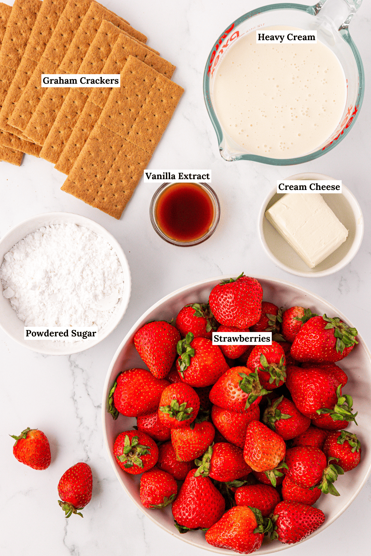 Top-down view of ingredients on a white surface: graham crackers, a bowl of strawberries, a block of cream cheese, a measuring cup of heavy cream, vanilla extract in a small bowl, and a bowl of powdered sugar.