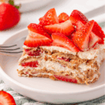 A slice of layered strawberry dessert with whipped cream and graham crackers, topped with fresh strawberry pieces, served on a white plate with a fork.