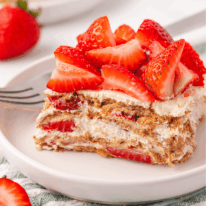 A slice of layered strawberry dessert with whipped cream and graham crackers, topped with fresh strawberry pieces, served on a white plate with a fork.