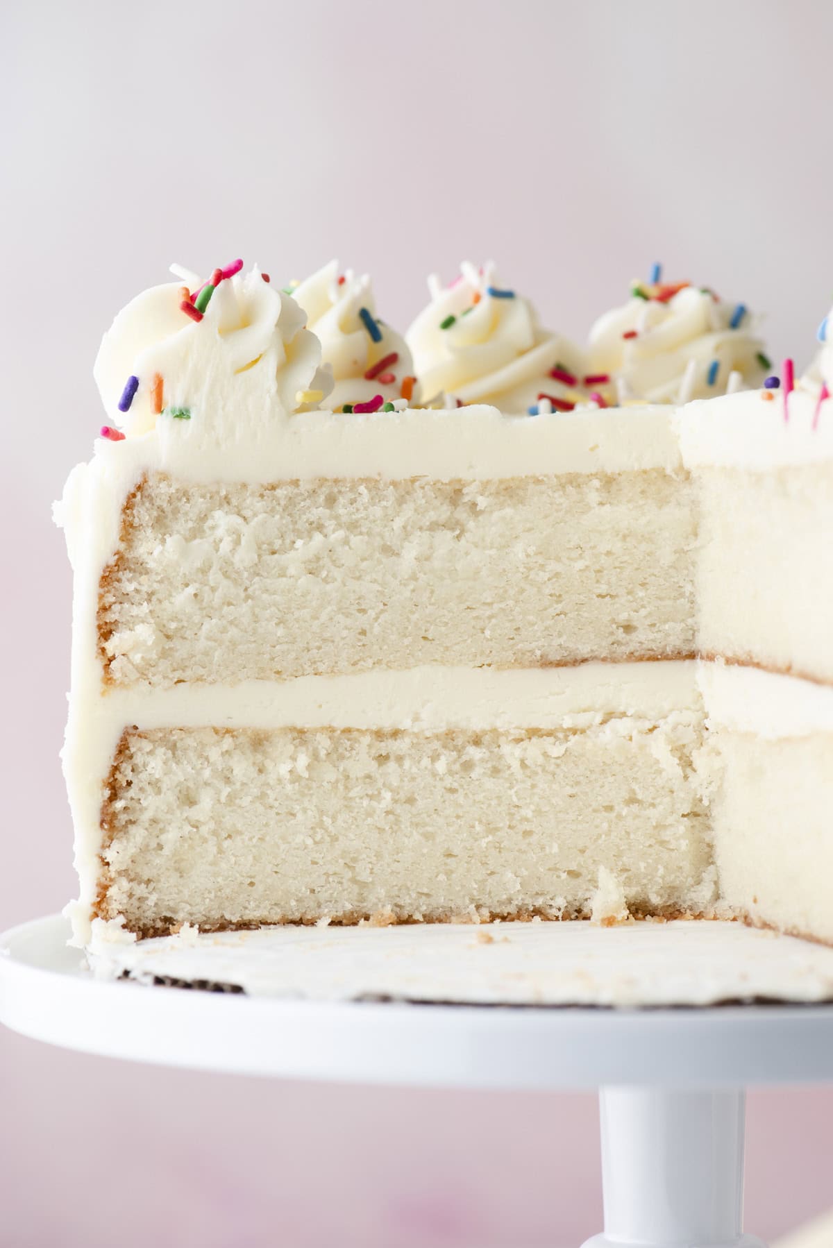 A close-up of a white frosted layer cake with two thick layers, topped with swirls of frosting and colorful rainbow sprinkles, on a white cake stand. A large slice has been cut out, revealing the cakes texture.