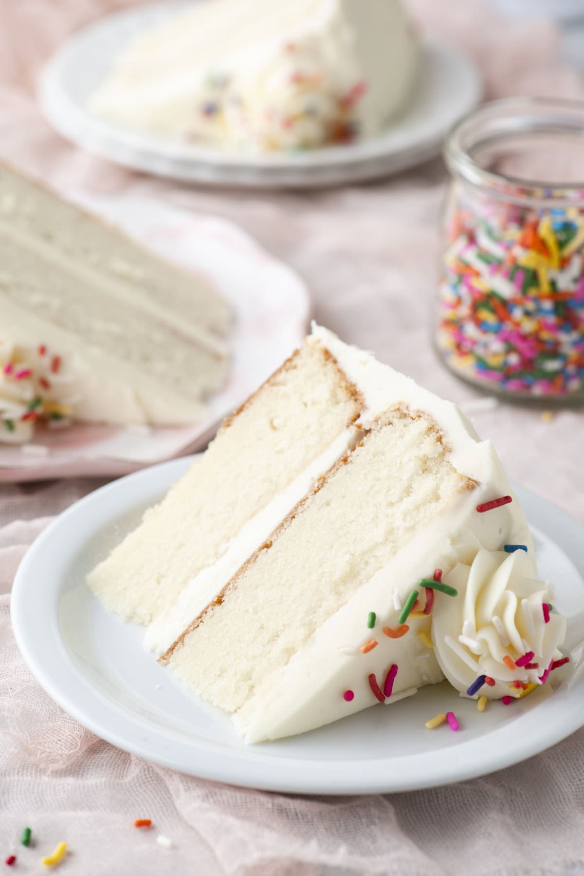 A slice of white layer cake with white frosting and colorful sprinkles sits on a white plate, with more cake slices and a jar of sprinkles in the background.