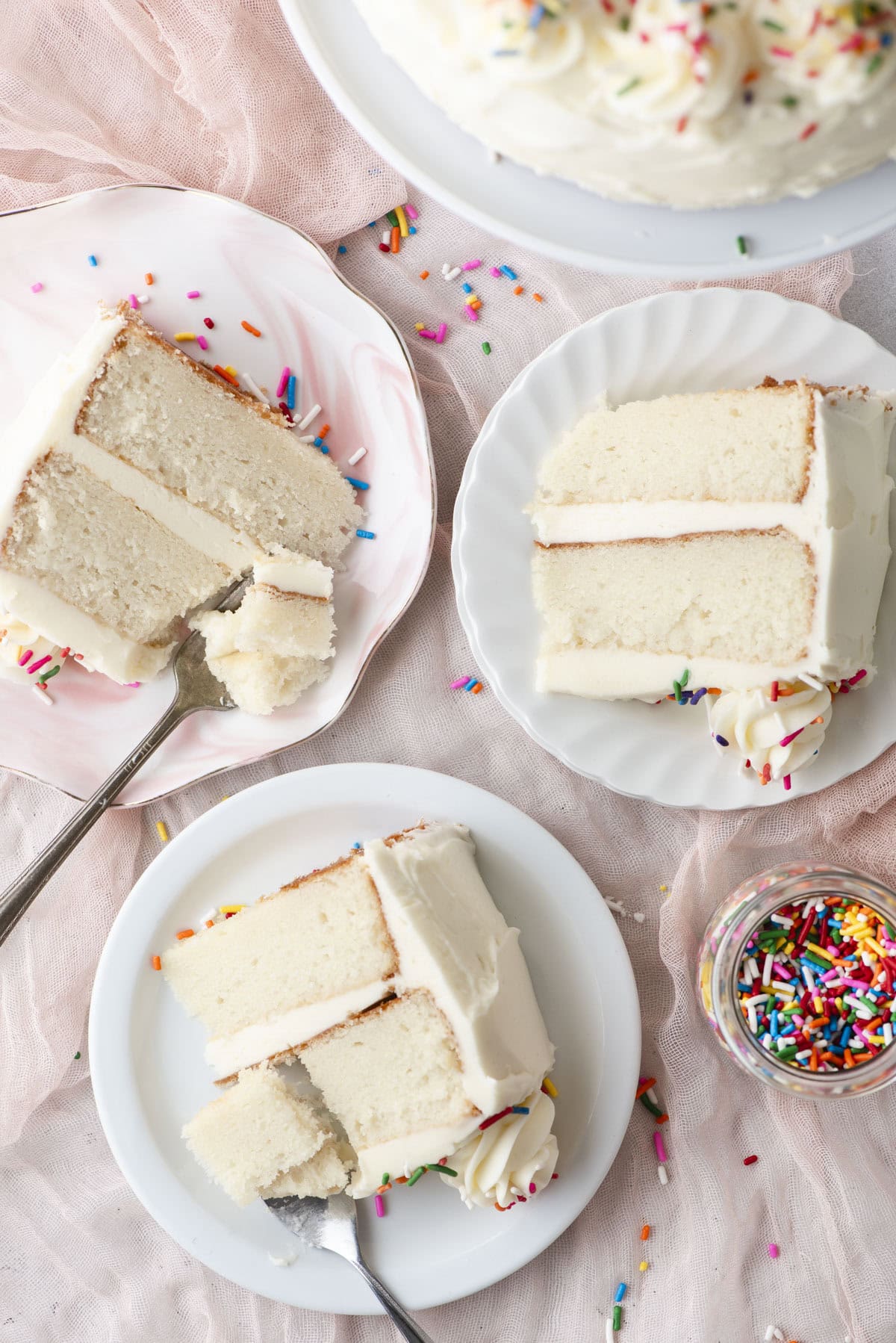 Three plates, each with a slice of white cake topped with white frosting and colorful sprinkles. A small jar of sprinkles and a light pink cloth are nearby, creating a festive dessert scene.
