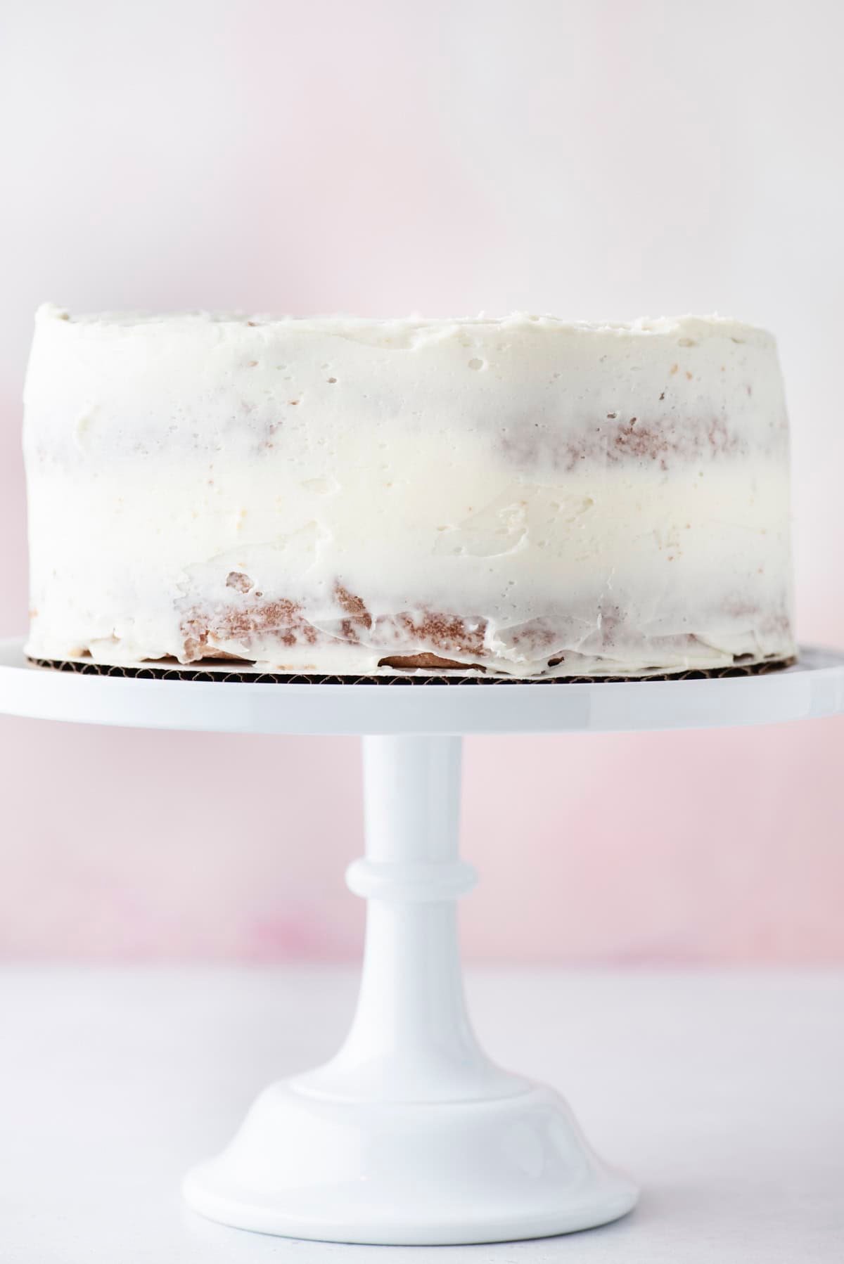 A simple, round cake with a thin layer of white frosting, partially revealing the cake underneath, sits on a white cake stand against a light pink and white background.