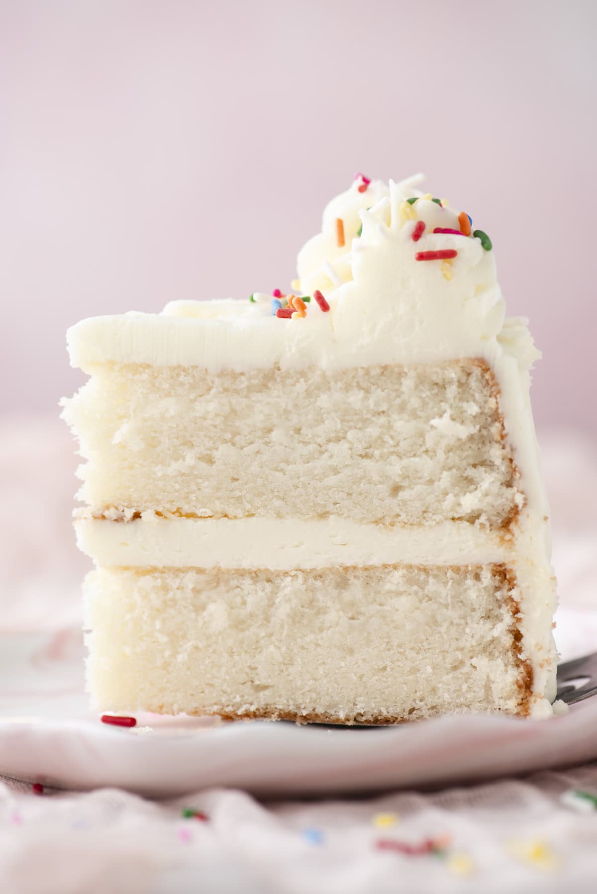 A close-up of a slice of white layer cake with white frosting, topped with colorful sprinkles, on a white plate against a soft pink background.
