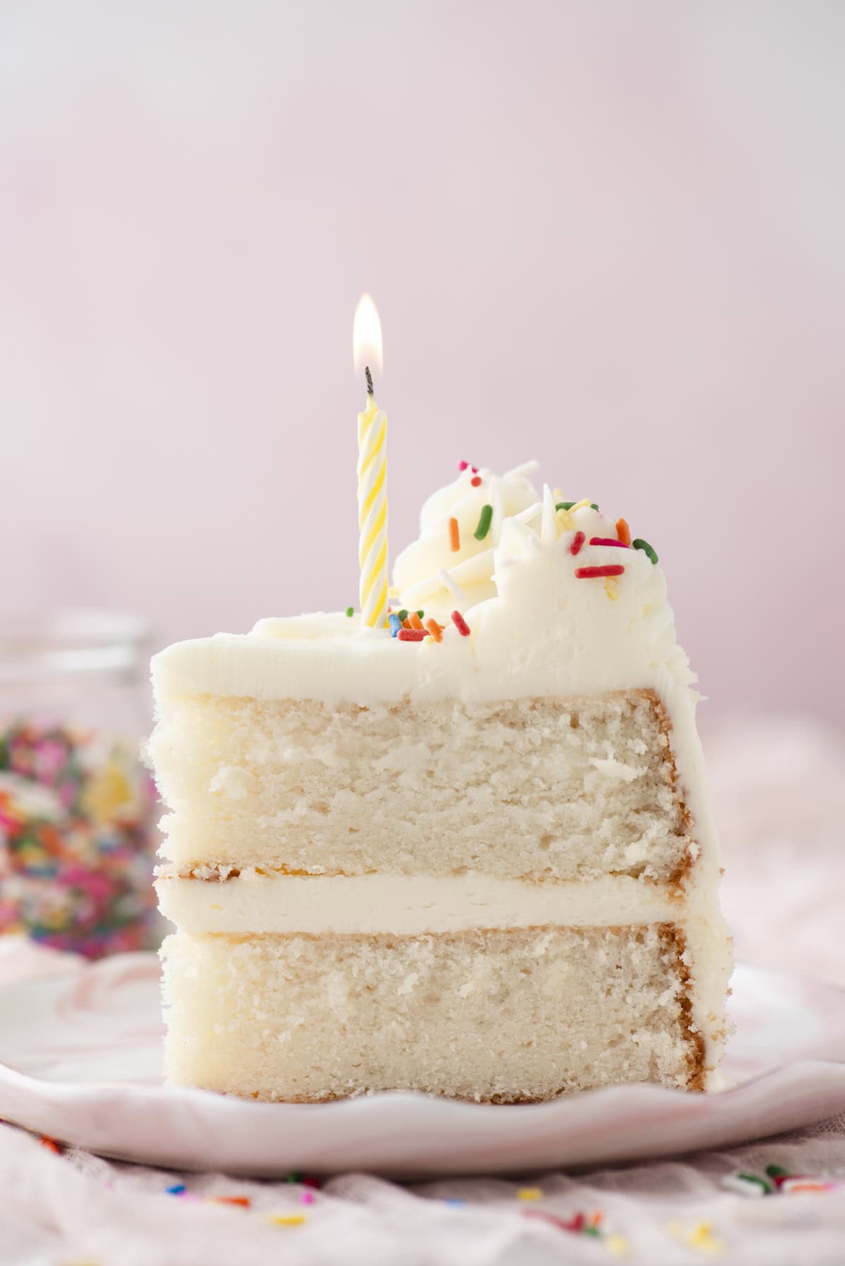 A slice of white layer cake with white frosting, rainbow sprinkles, and a lit yellow-striped candle on top sits on a plate. The background is softly blurred with a jar of sprinkles visible.