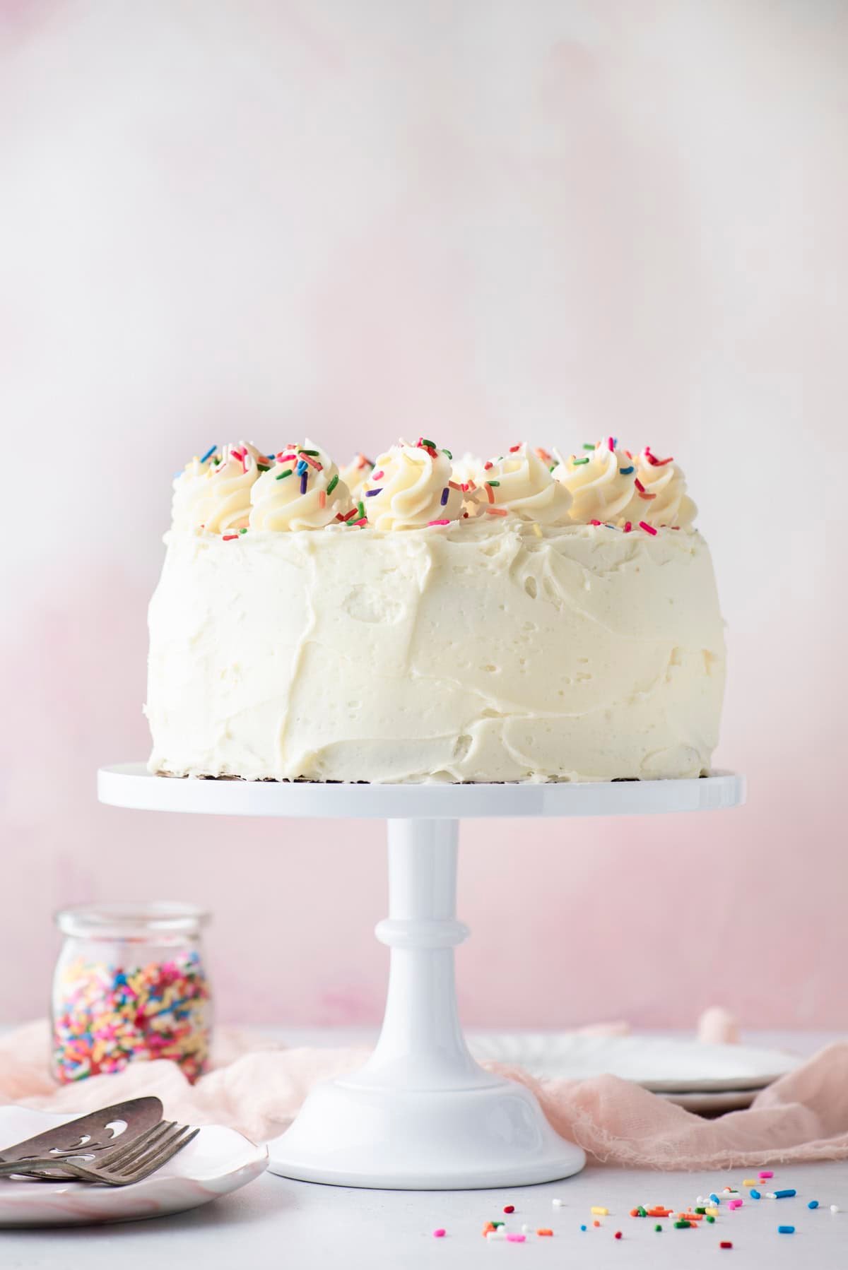 A round white frosted cake topped with swirls of frosting and colorful sprinkles sits on a white cake stand. A jar of sprinkles and a cake server are on the table nearby. The background is soft pink.